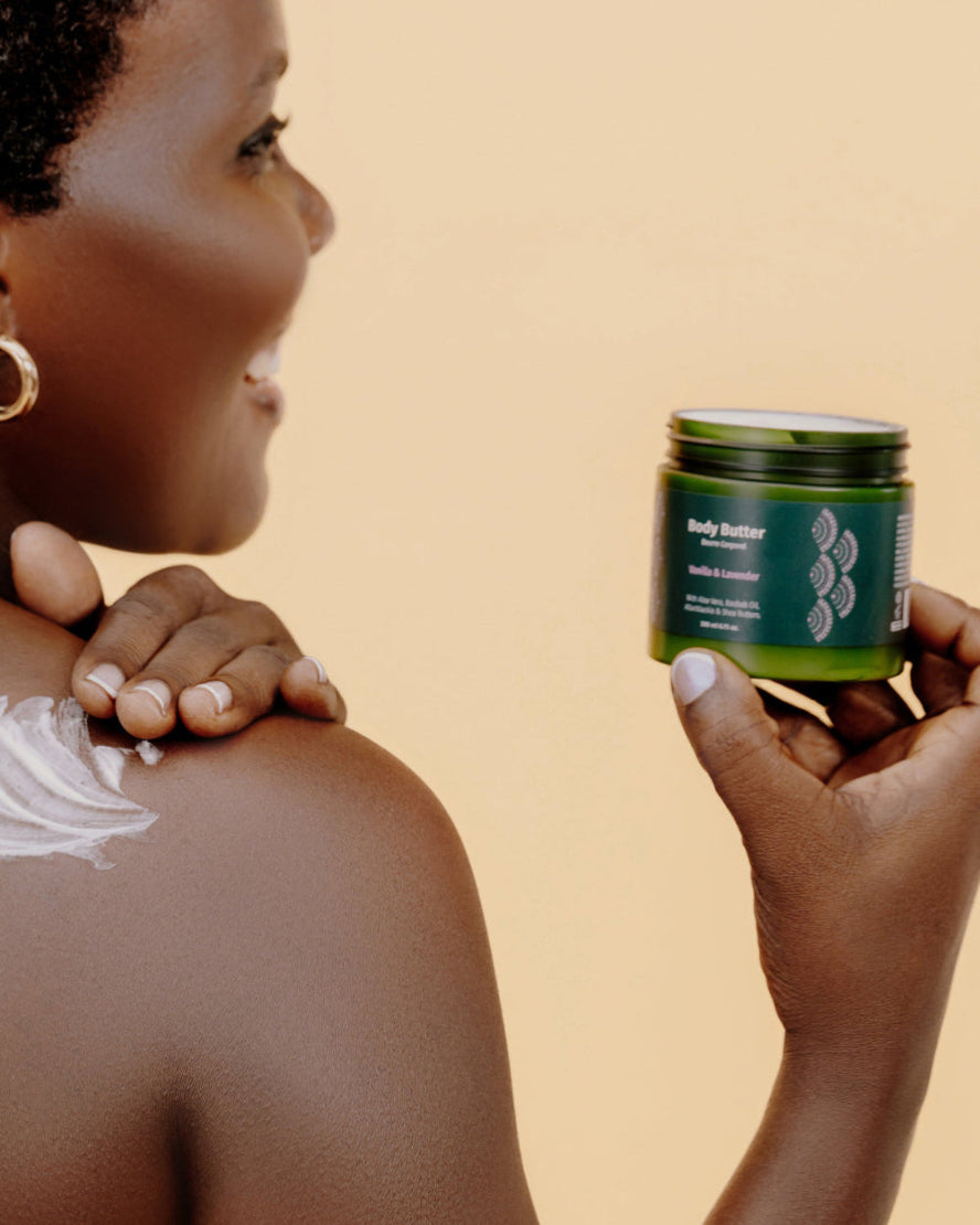 African woman applies cream to her shoulder while holding a green jar of Body Butter.